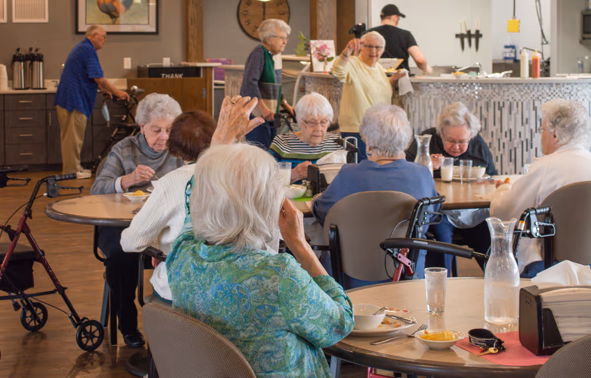A group of elderly people sitting around tables in a dining area, eating and interacting. Some use walkers and wheelchairs. In the background, a staff member is working behind a counter, and another elderly person is standing and waving.