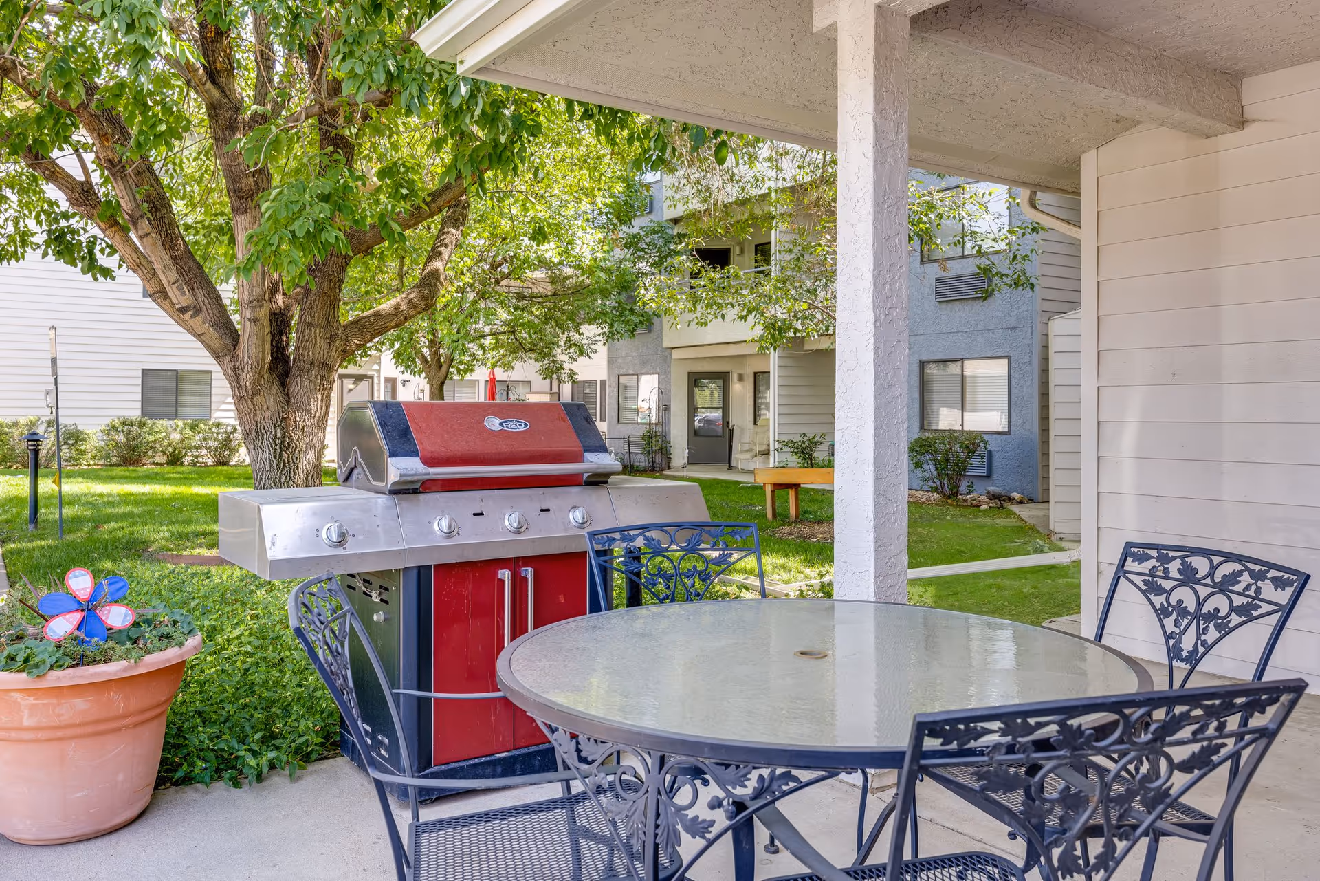Outdoor patio area at Greeley Place featuring a round glass-top table with four ornate metal chairs, a large stainless steel and red gas grill, a potted plant with a colorful pinwheel, and green trees and grass surrounding the space with apartment buildings in the background.