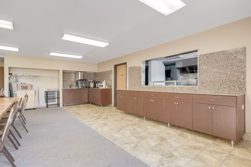 Interior room with a long counter and cabinets along the right wall, a serving window into a kitchen area, and a coffee station in the corner. There are tables and chairs on the left side of the room, with a carpeted floor transitioning to tiled flooring near the counter. The room is well-lit with ceiling lights.