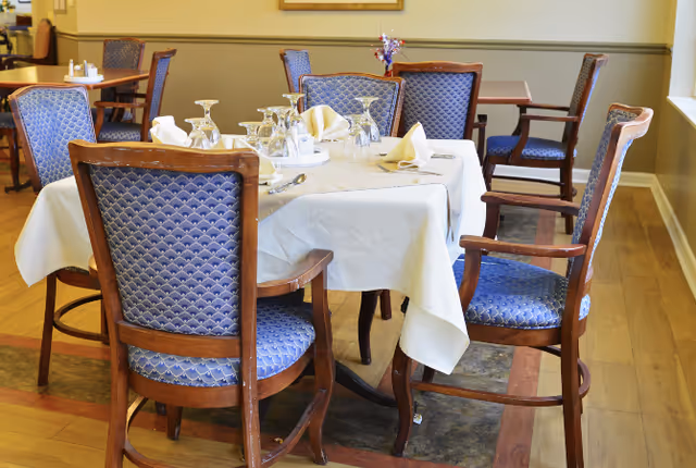 A dining table set for a meal with a white tablecloth, folded napkins, upside-down wine glasses, and a small floral centerpiece. The table is surrounded by wooden chairs with blue patterned upholstery. Additional tables and chairs are visible in the background in a well-lit room with wooden flooring.