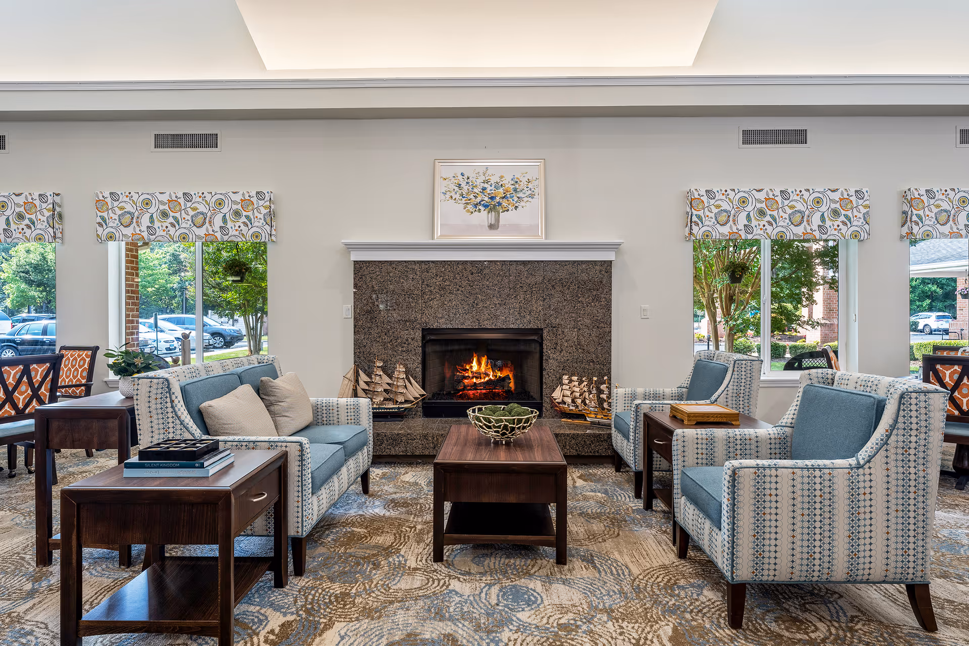 A cozy living room area in Colonial Harbor Gracious Retirement Living featuring a lit fireplace with a granite surround. The room has two patterned armchairs and a matching loveseat with blue cushions arranged around a wooden coffee table. There are two wooden side tables with books and decorative items. Large windows with floral valances let in natural light and show a view of parked cars and greenery outside. A framed floral painting hangs above the fireplace.