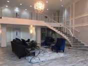 Spacious modern lobby with sofas and chairs arranged on a rug, a decorative chandelier, and a curved staircase leading to a mezzanine.