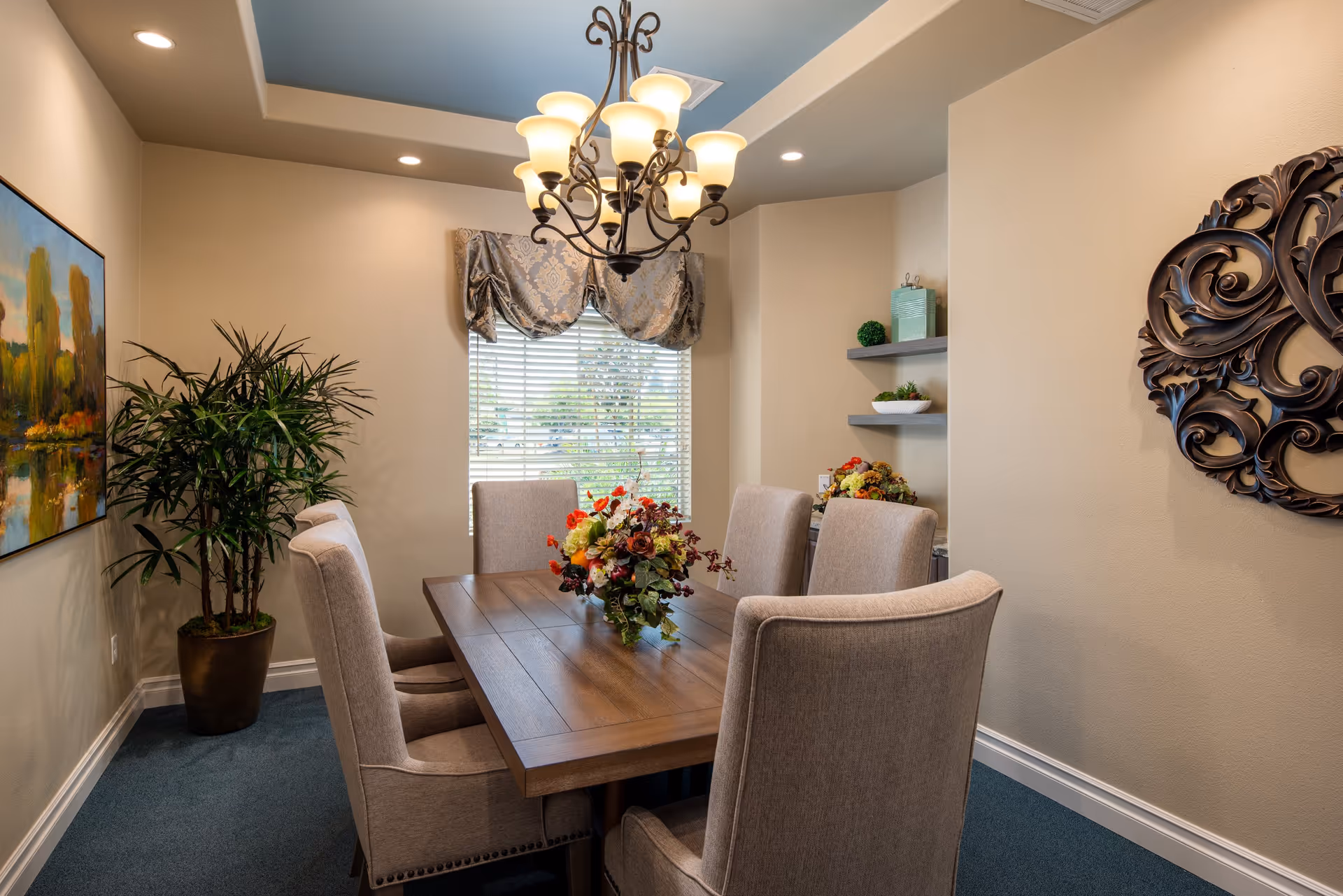 A cozy dining room with a wooden table surrounded by six beige upholstered chairs. A floral centerpiece decorates the table. The room features a window with blinds and a decorative valance, a chandelier with multiple lights hanging from the ceiling, a large potted plant in the corner, a colorful landscape painting on one wall, and decorative shelves with small plants and items on another wall.