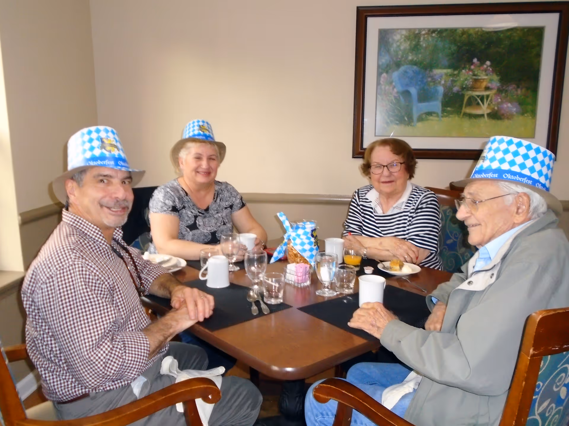 Four seniors wearing blue-and-white Oktoberfest hats seated around a dining table with drinks and snacks.
