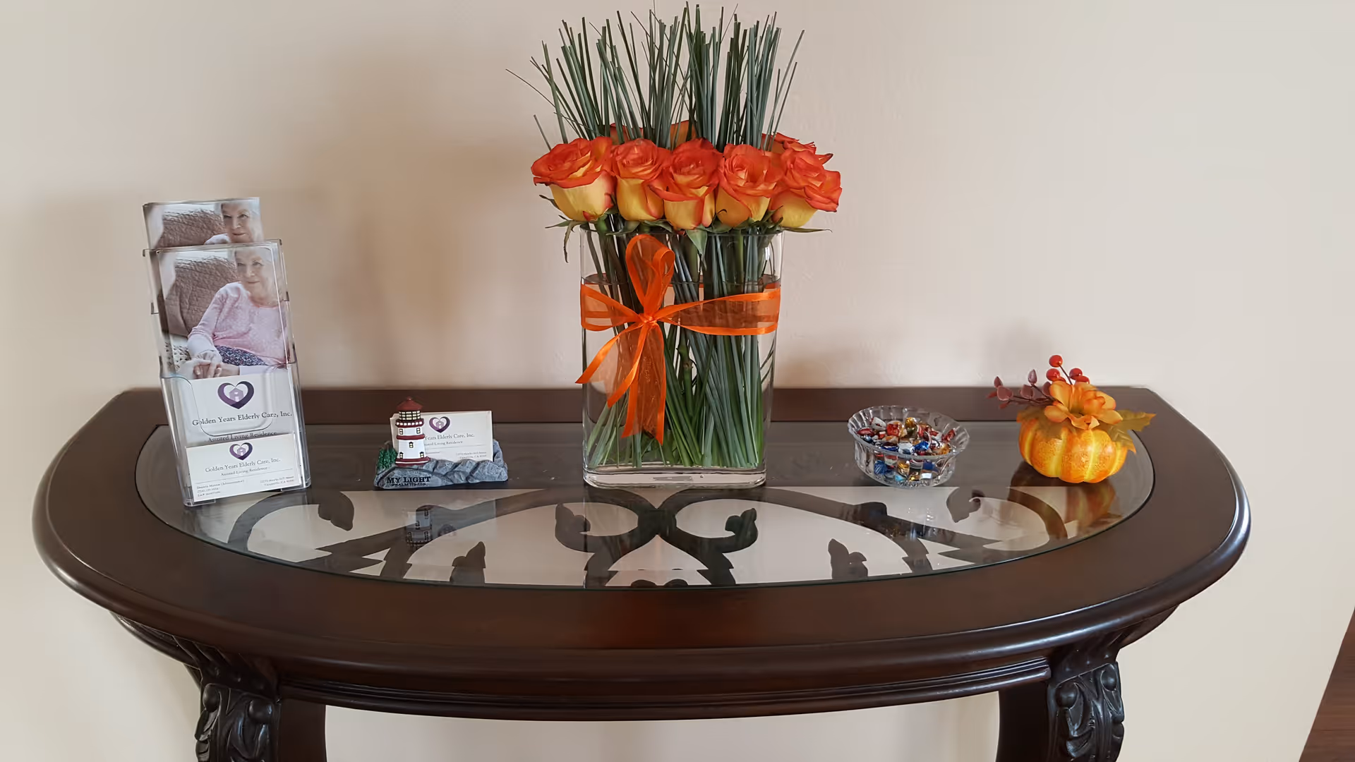 A dark wooden console table with a glass top against a beige wall. On the table, there is a clear vase with orange roses tied with an orange ribbon, a small bowl of assorted candies, a small decorative pumpkin with autumn leaves, and a brochure holder containing brochures for Golden Years Elderly Care, Inc.