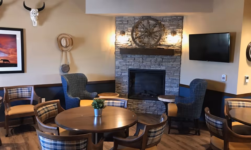 Cozy senior living common room with round wooden tables, plaid chairs, armchairs, and a stone fireplace topped by a decorative wagon wheel.