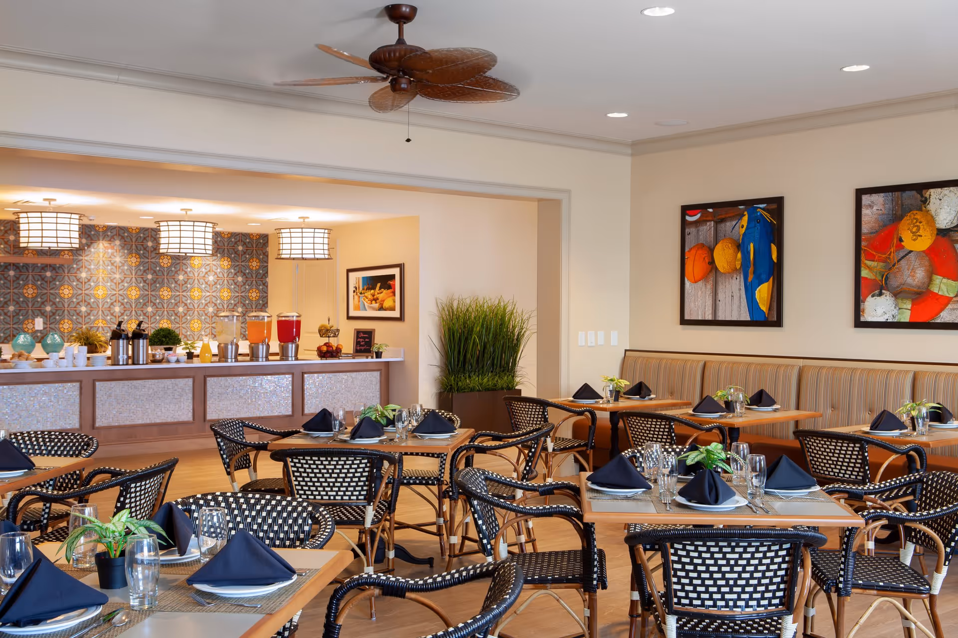 A bright dining room with multiple tables set with plates, glasses, and black napkins. The chairs have a woven black and white pattern. In the background, there is a buffet area with beverage dispensers and decorative items, and the wall behind it features patterned tiles. Two colorful framed artworks hang on the beige walls, and a ceiling fan with leaf-shaped blades is visible above.