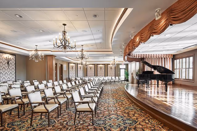 A spacious, elegant event room with rows of white cushioned chairs arranged facing a wooden stage. On the stage, there is a black grand piano. The room features ornate chandeliers hanging from the ceiling, patterned carpet, and large windows with blinds allowing natural light to enter.
