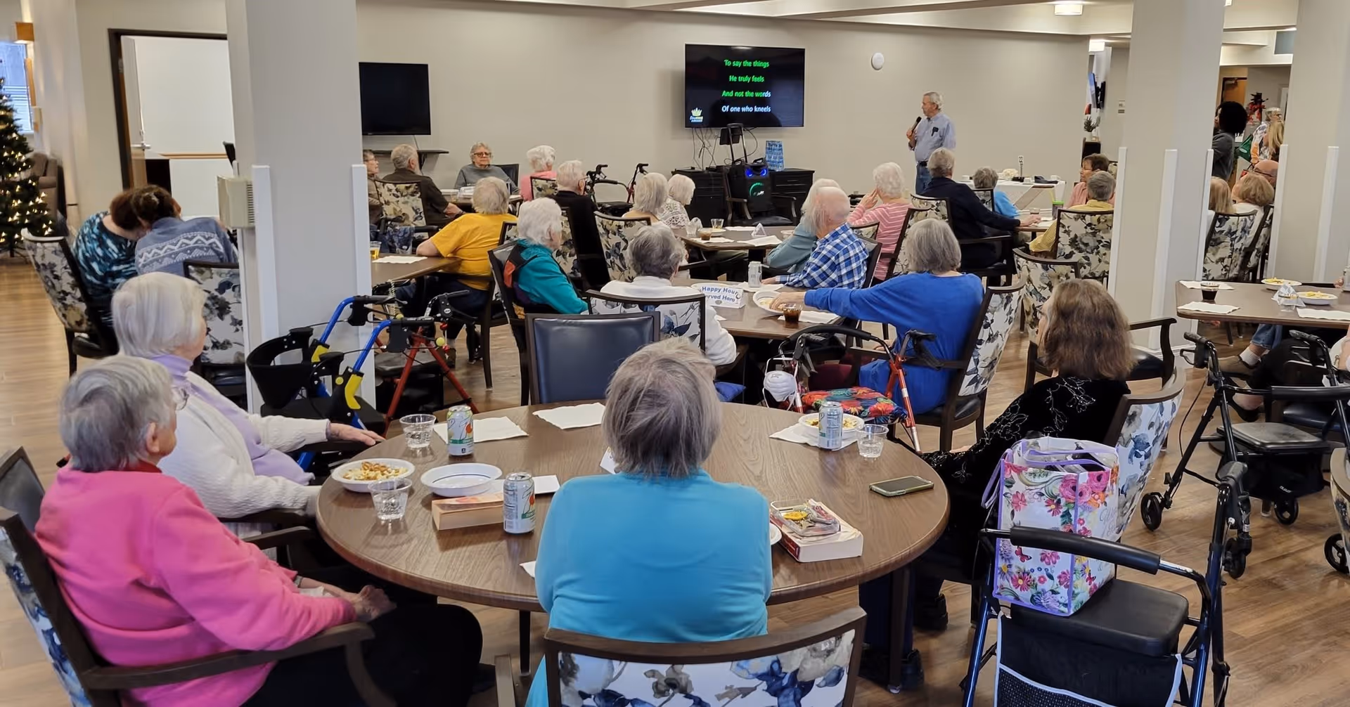 A group of elderly residents seated around multiple tables in a senior living facility common area, attentively watching and listening to a man speaking at the front near a television screen displaying karaoke lyrics. Several walkers and wheelchairs are visible, and the room has a warm, communal atmosphere.