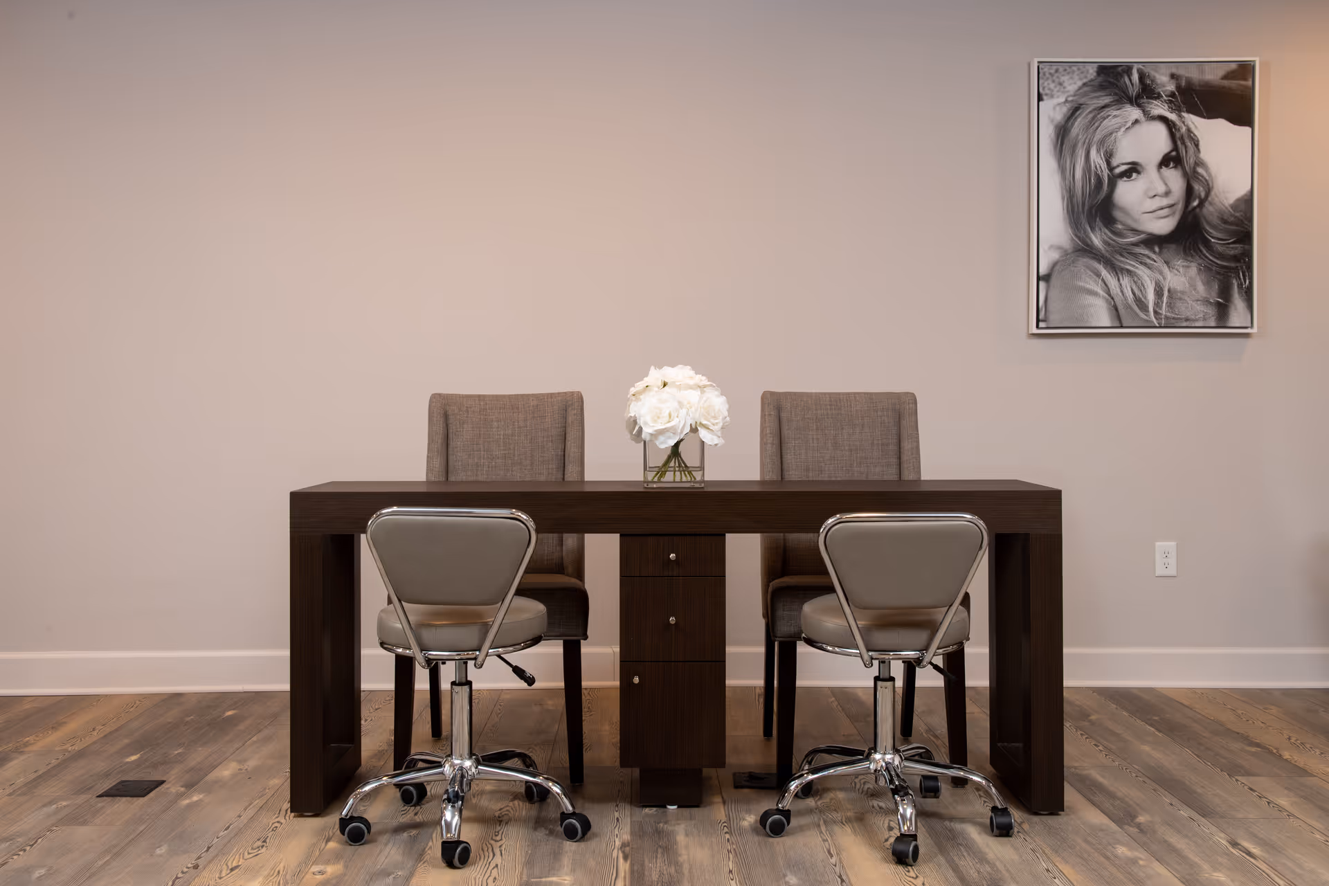 A minimalist interior with a dark wood desk, two rolling chairs, two upholstered chairs, a vase of white flowers, and a black-and-white portrait on the wall.