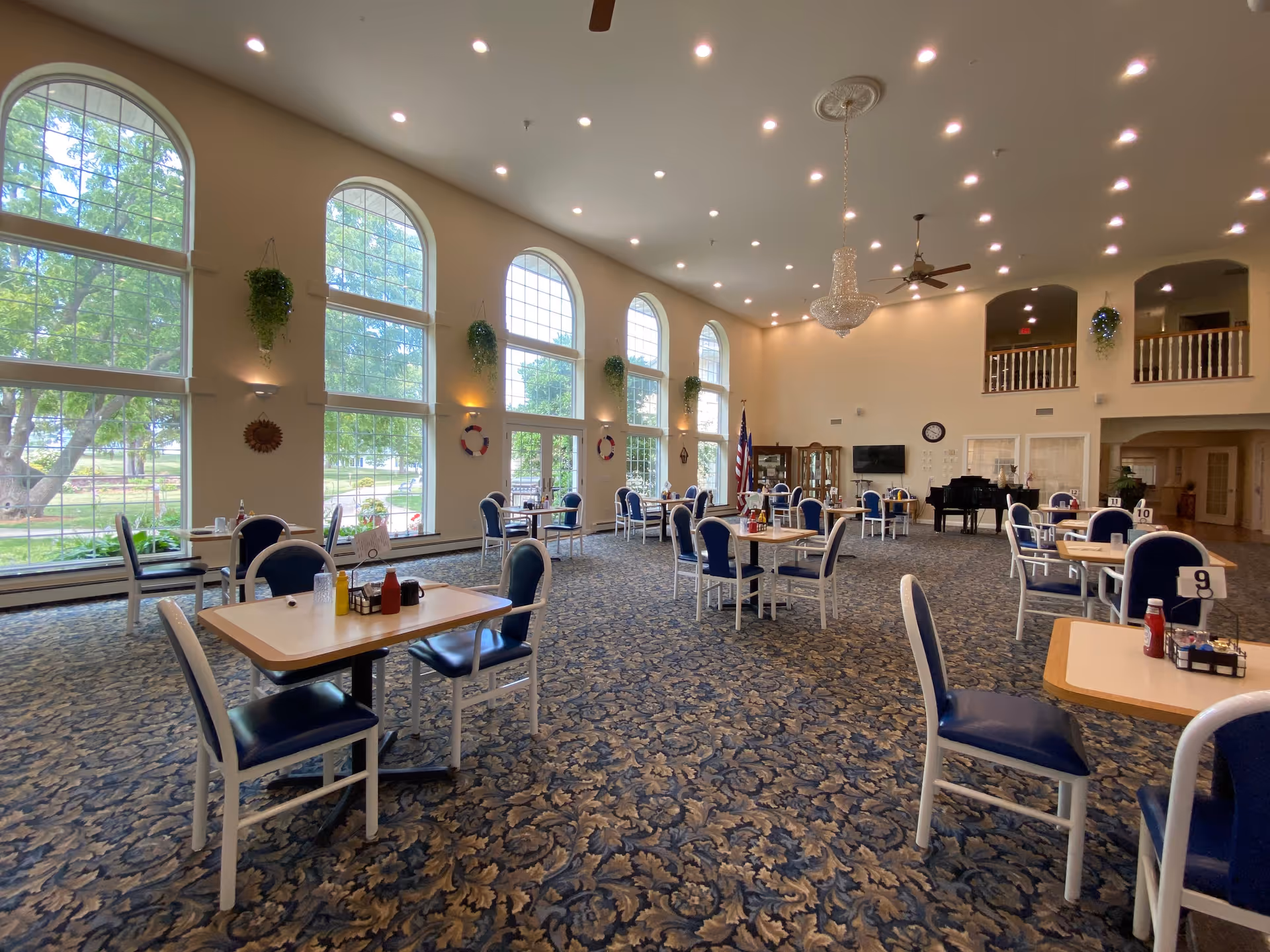 Spacious dining room with multiple tables and chairs arranged neatly. Large arched windows allow natural light to fill the room, with hanging plants between the windows. The room features a patterned carpet, a chandelier, ceiling fans, and an American flag near a piano and a display cabinet.
