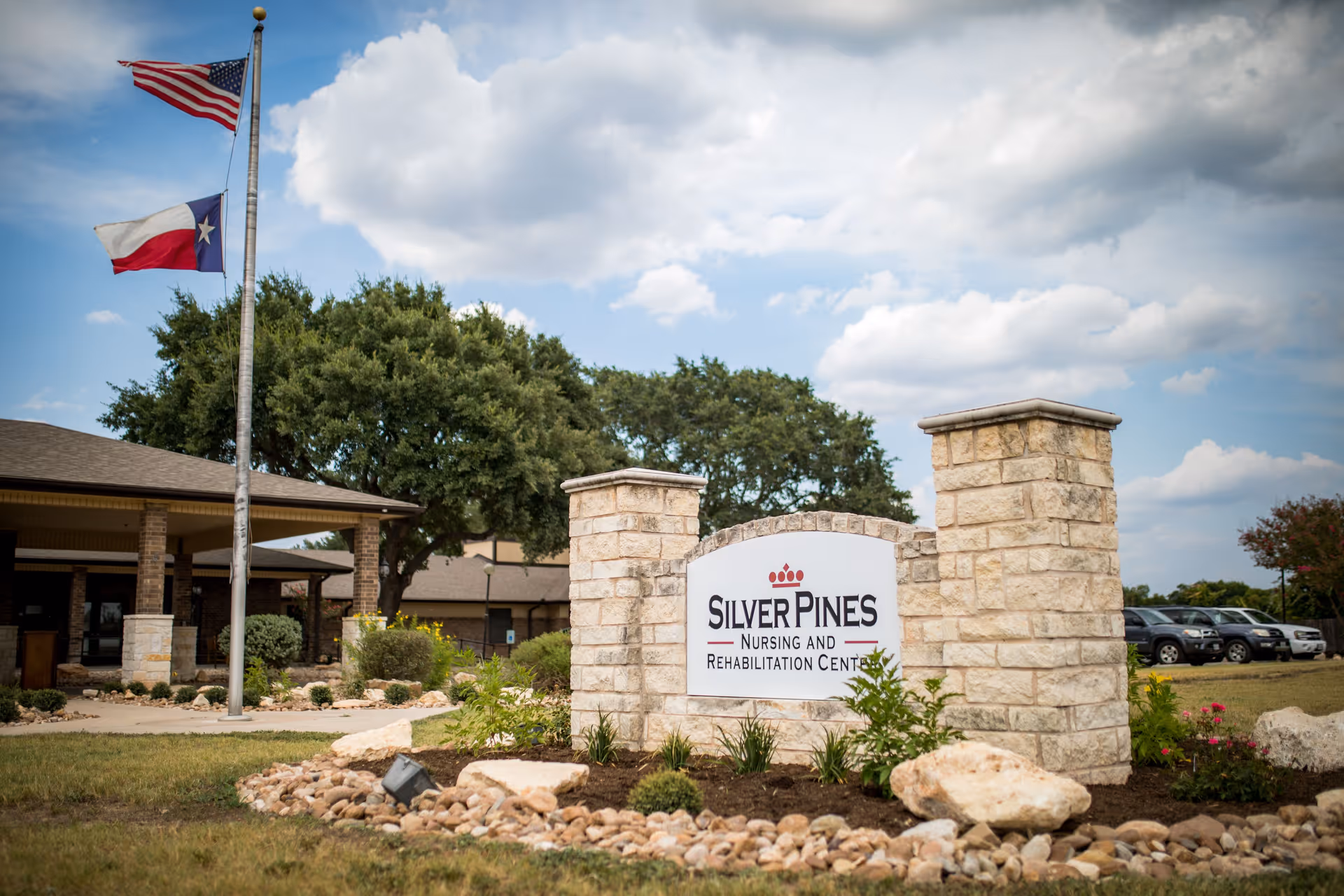 Exterior view of Silver Pines Nursing and Rehabilitation Center with a stone sign in the foreground, two flagpoles flying the American and Texas flags, and a building with a covered entrance in the background under a partly cloudy sky.