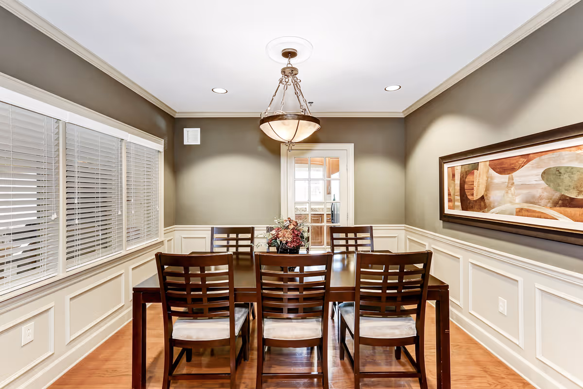 A dining room with a dark wooden table and six matching chairs with cushioned seats. A floral centerpiece is placed on the table. The walls are painted gray with white wainscoting, and a large abstract painting hangs on the right wall. There are three windows with white blinds on the left wall and a glass-paneled door at the back. A decorative ceiling light fixture hangs above the table, and the floor is wooden.