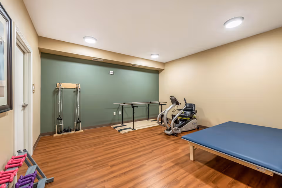 Therapy/exercise room with parallel bars, a recumbent exercise bike, a treatment table and free weights on wood flooring.