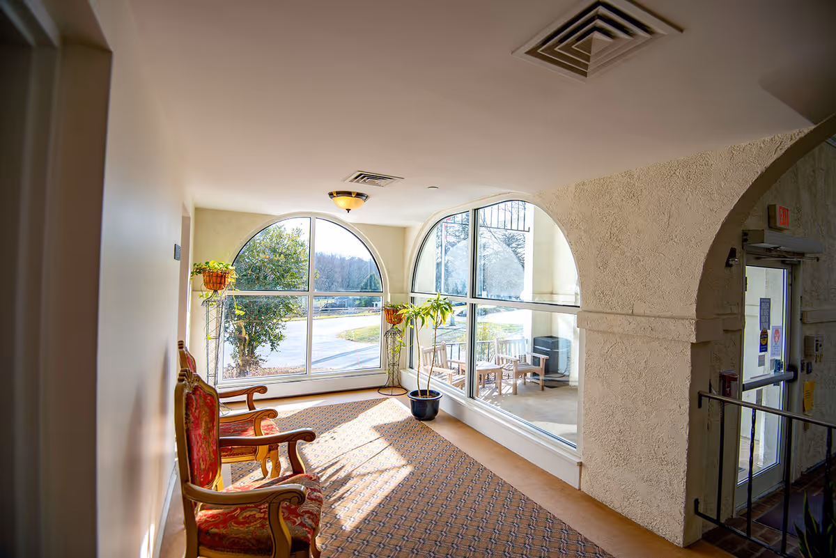 Sunlit seating alcove in a nursing facility with two upholstered chairs, potted plants, and large arched windows overlooking the exterior.