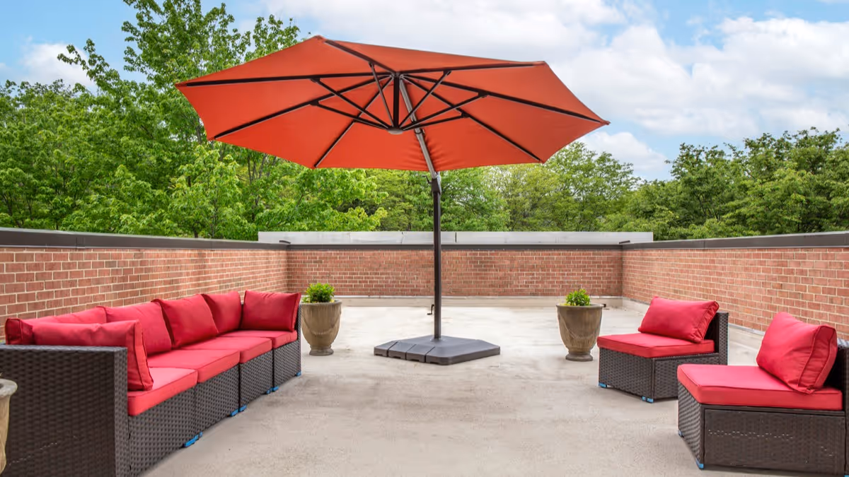 Outdoor patio area with a large red umbrella in the center, surrounded by dark wicker furniture with red cushions, and two large planters with greenery against a backdrop of brick walls and trees.
