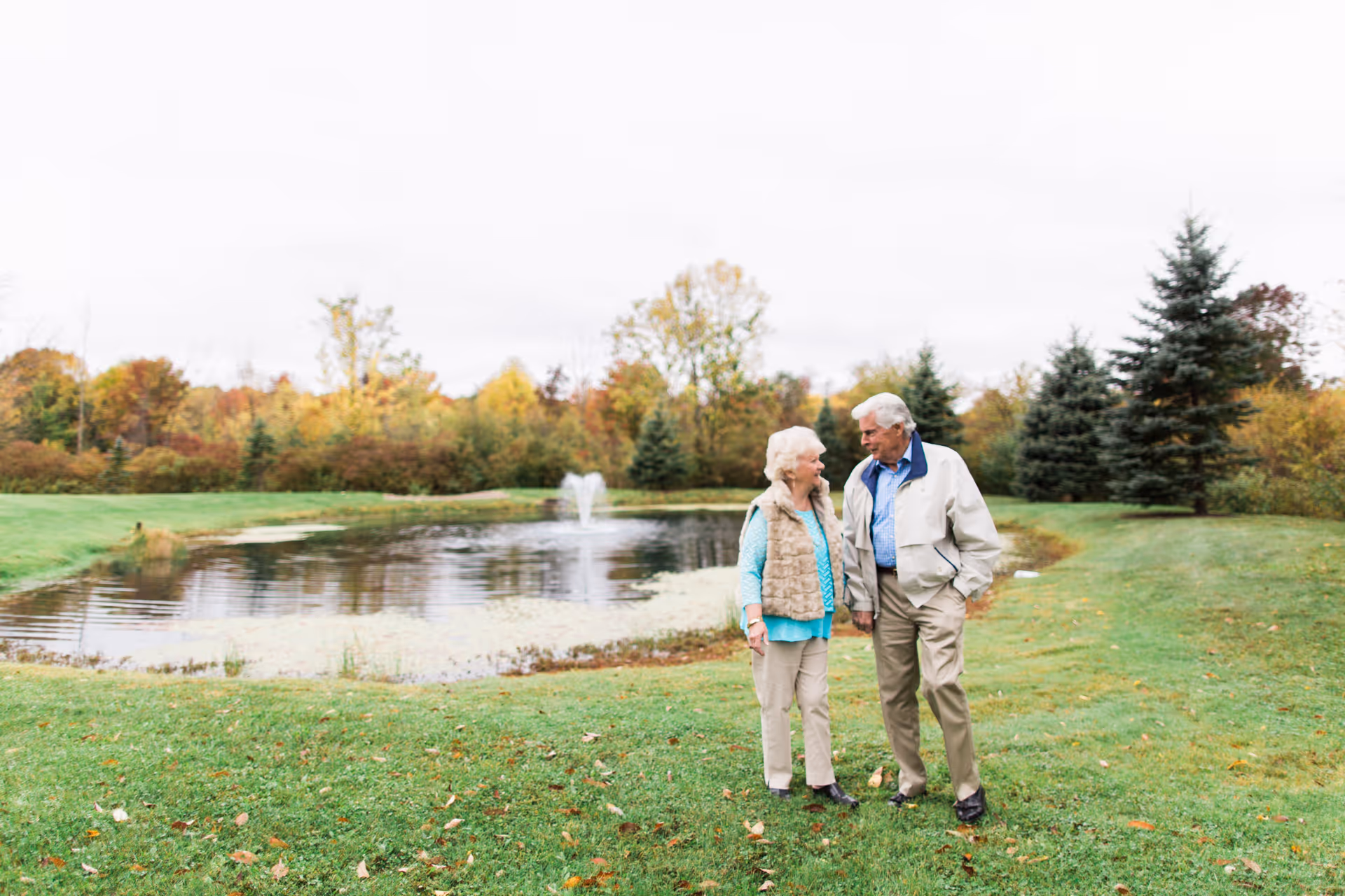 An elderly couple walking and talking beside a small pond with a fountain in a grassy outdoor area surrounded by trees with autumn foliage.