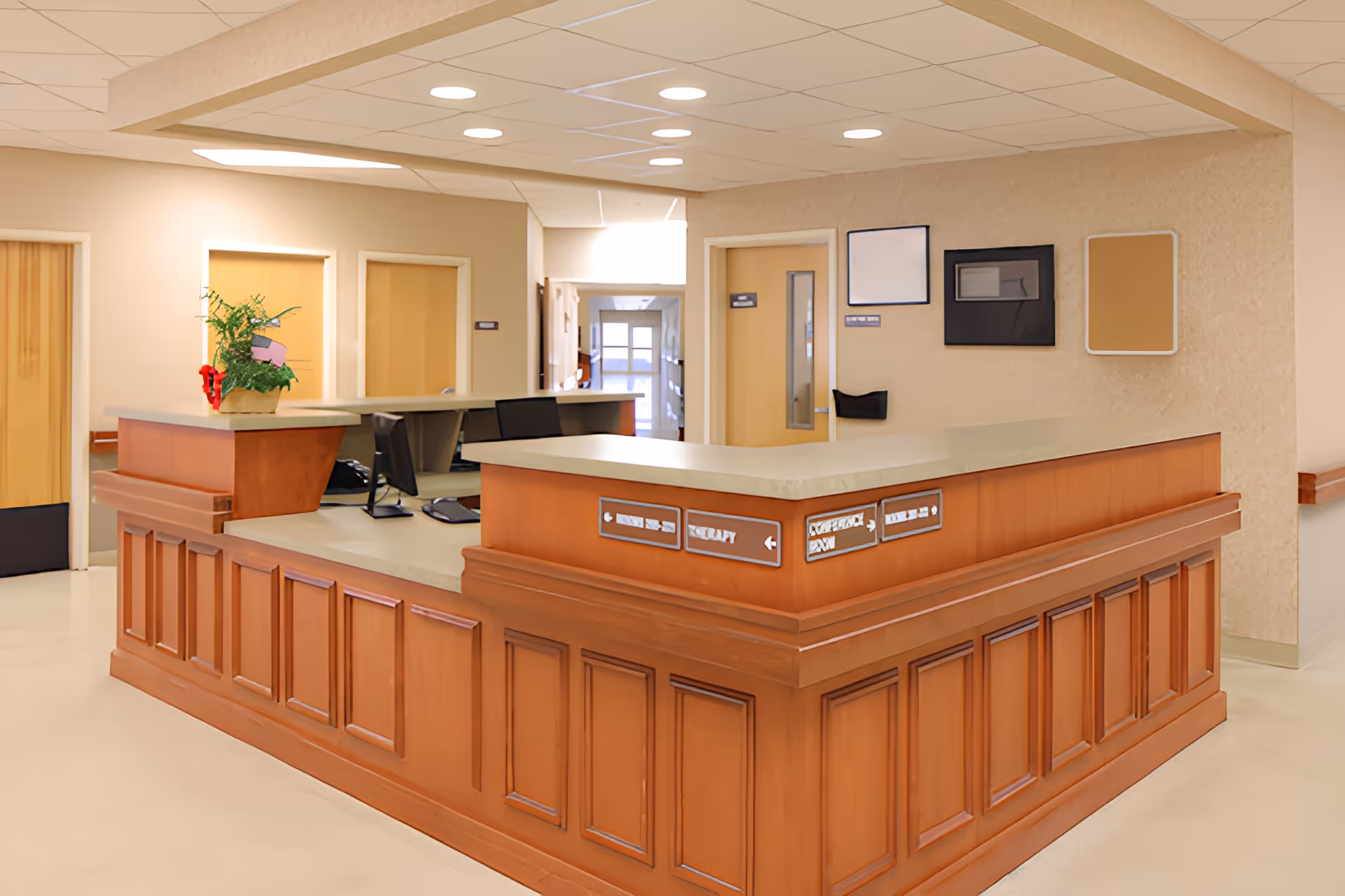 A reception desk area inside a senior living facility with a wooden counter and beige walls. There are computer monitors on the desk, a potted plant with red flowers, and several doors and bulletin boards on the walls in the background. The ceiling has recessed lighting.