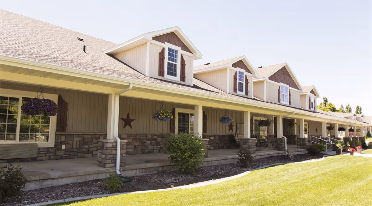 Exterior view of a single-story assisted living facility with a long covered porch, stone and wood siding, hanging flower baskets, and a well-maintained lawn in front.