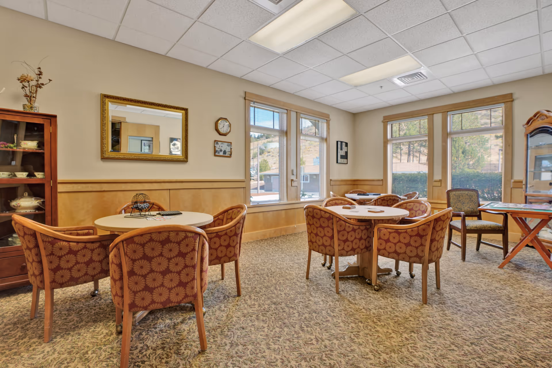 A well-lit common area with round tables and patterned chairs arranged for group activities or dining. The room features large windows with views of trees outside, a wooden cabinet displaying dishes, a wall mirror, and a clock. The carpet has a floral pattern and the walls are painted beige with wooden trim.