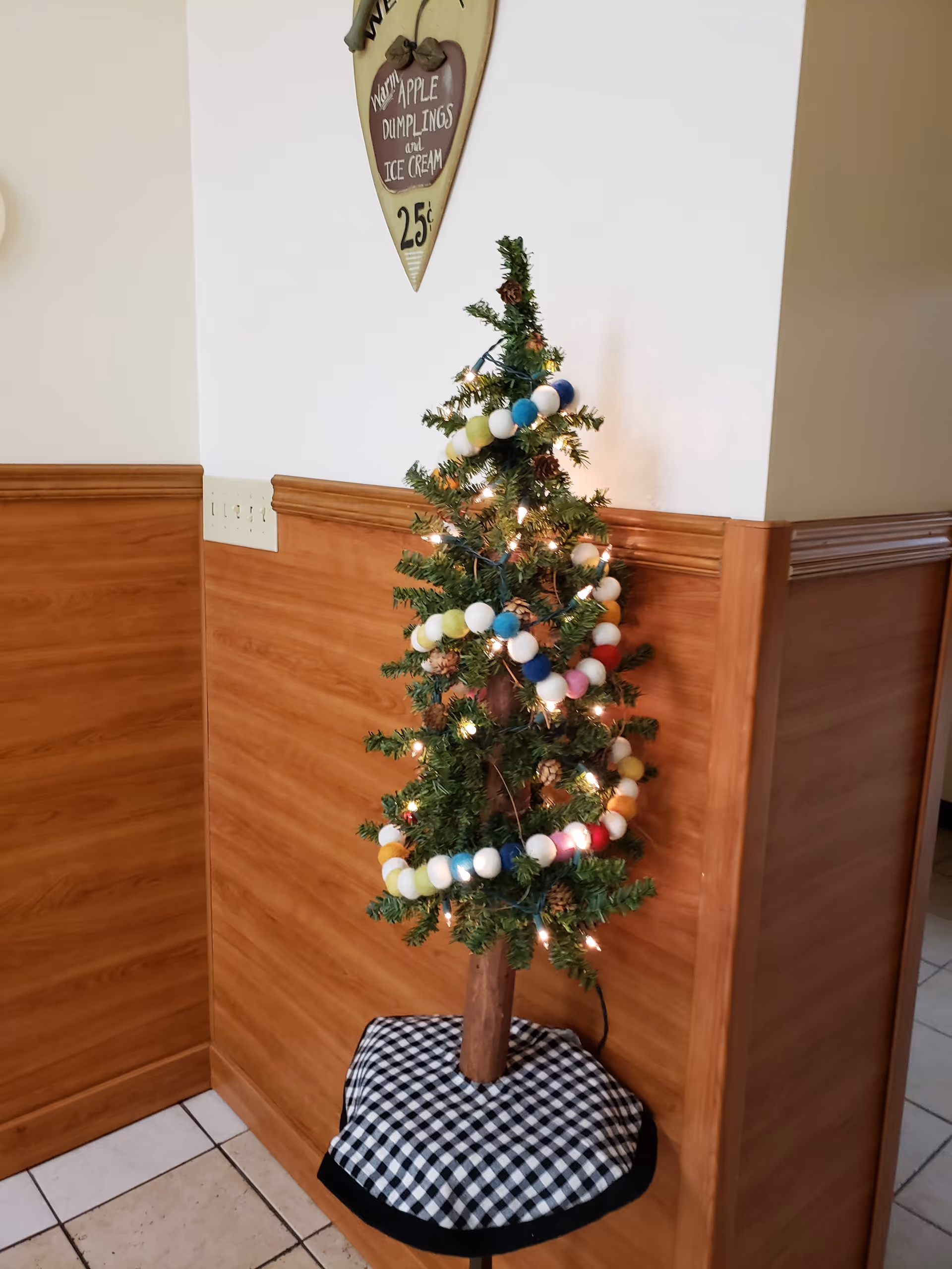 Small decorated Christmas tree with colorful pom-pom garland and lights in a wood-paneled corner of an interior hallway.