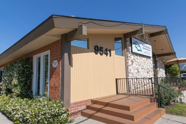 Exterior view of Panorama Gardens Nursing & Rehabilitation Center building with address number 9541 displayed on the front. The building features a combination of brick and stone walls, a small staircase leading to the entrance, and greenery along the side.