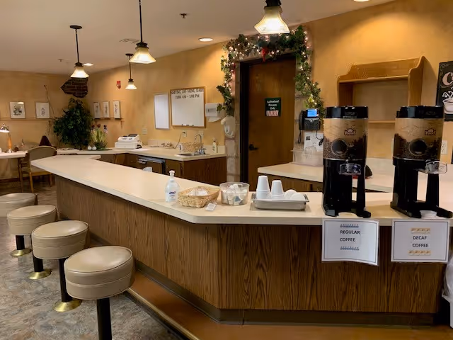 Interior view of a coffee and snack counter area in a senior living facility. The counter has stools, two coffee dispensers labeled Regular Coffee and Decaf Coffee, a basket with sugar packets, disposable cups, and a hand sanitizer bottle. The walls are decorated with framed pictures, a plant, and a door adorned with a garland. The lighting is warm with hanging pendant lights.