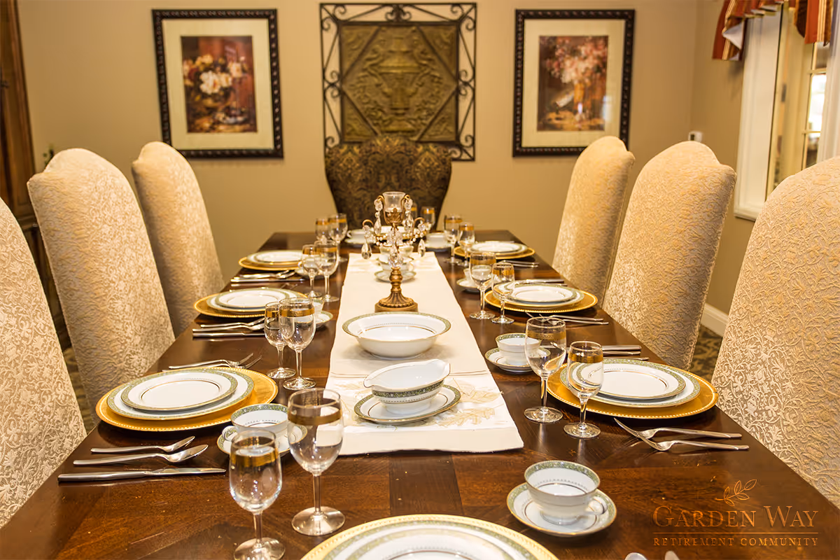 A formal dining room with a long wooden table set for a meal. The table is adorned with a white runner, gold-rimmed plates, bowls, cups, and multiple glasses at each place setting. The chairs are upholstered in a light patterned fabric. The walls have framed floral artwork and decorative metal wall art. Natural light comes through a window on the right side.