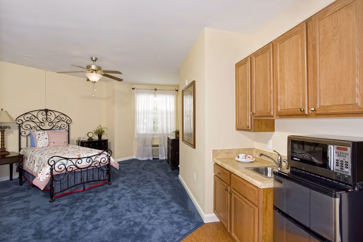 A senior living facility room featuring a single bed with a decorative metal headboard and floral bedding, a nightstand with a lamp and clock, a window with sheer curtains, and a kitchenette area with wooden cabinets, a small sink, a microwave, and a mini refrigerator.