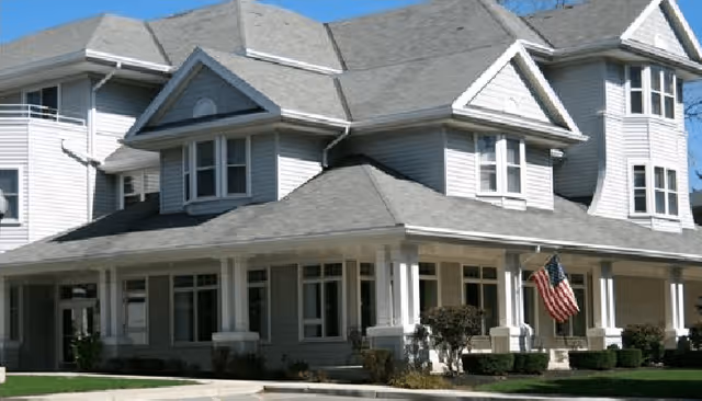Exterior view of a large, multi-story residential building with gray siding and a gray roof. The building features multiple windows, a covered porch with white columns, and an American flag displayed near the entrance. The sky is clear and blue.