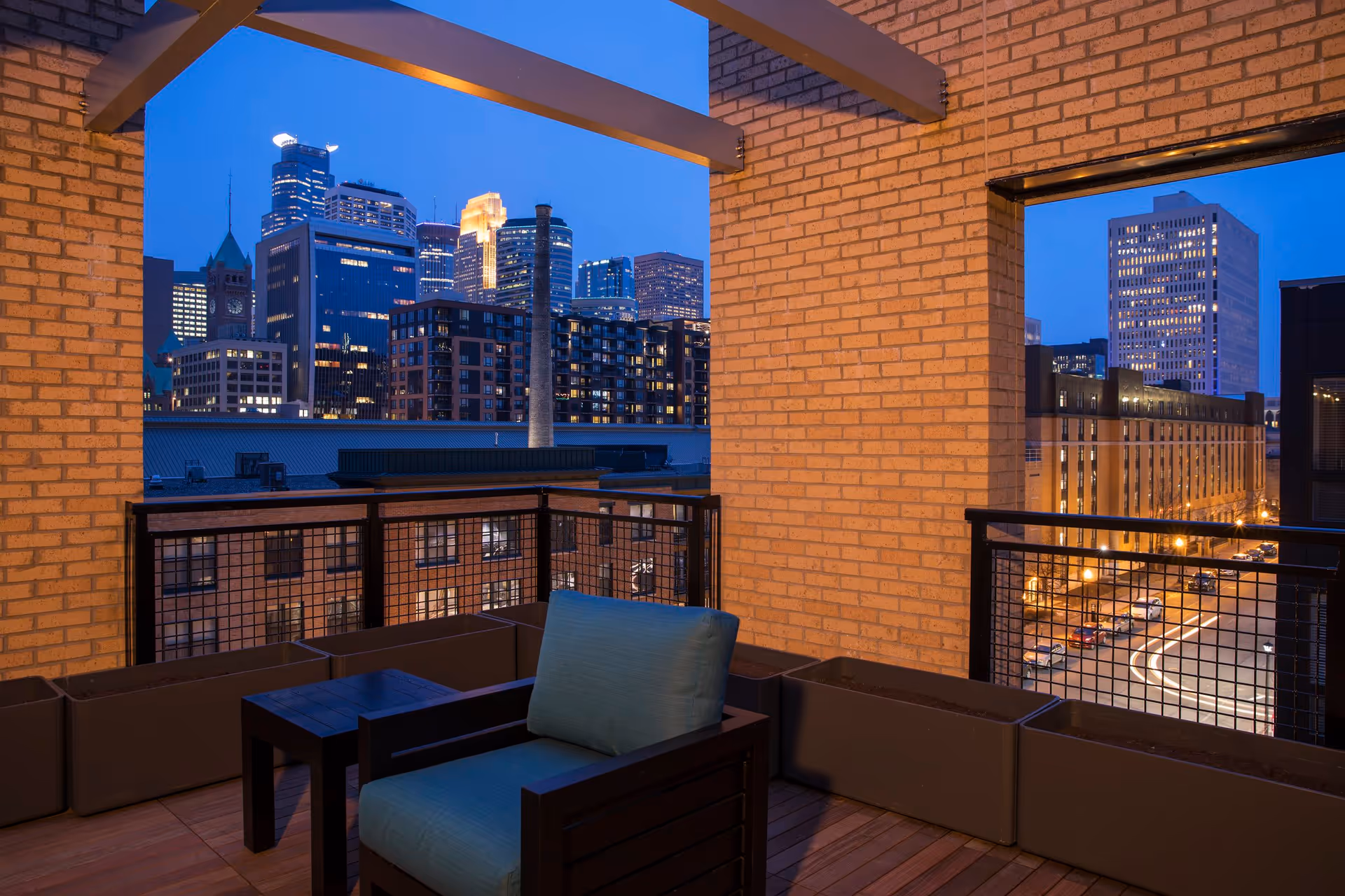 View from a balcony at dusk showing a comfortable chair with a blue cushion and a small table, framed by brick walls and railings, overlooking a cityscape with illuminated buildings and streets.