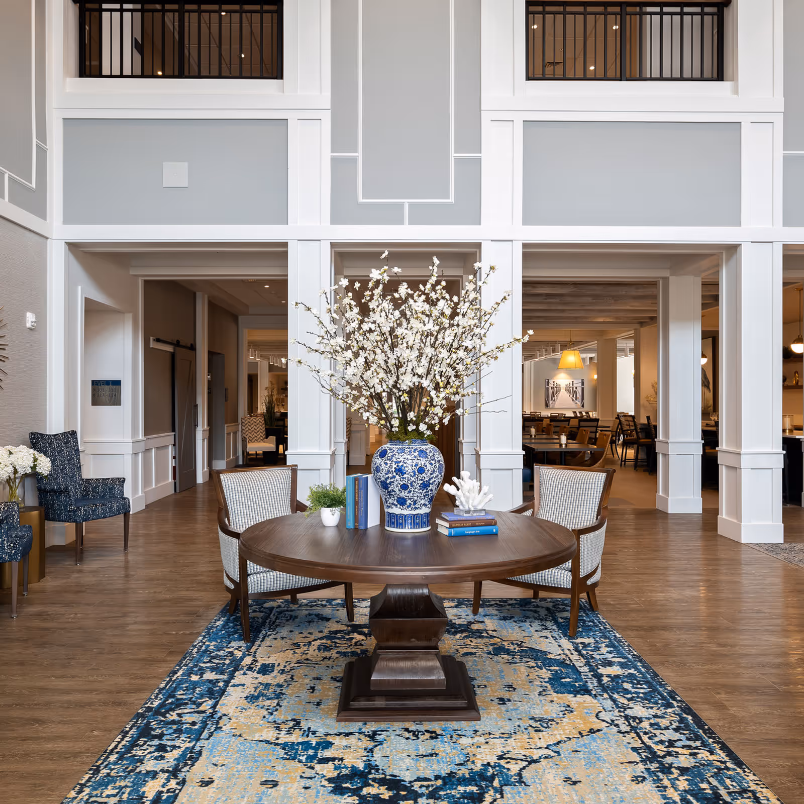 A spacious and elegant senior living facility common area with a round wooden table at the center, decorated with a large blue and white vase filled with white flowers, books, and a small plant. The room features high ceilings, white paneled walls, wood flooring, and a blue and beige patterned rug under the table. There are two upholstered chairs around the table and additional seating visible in the background.