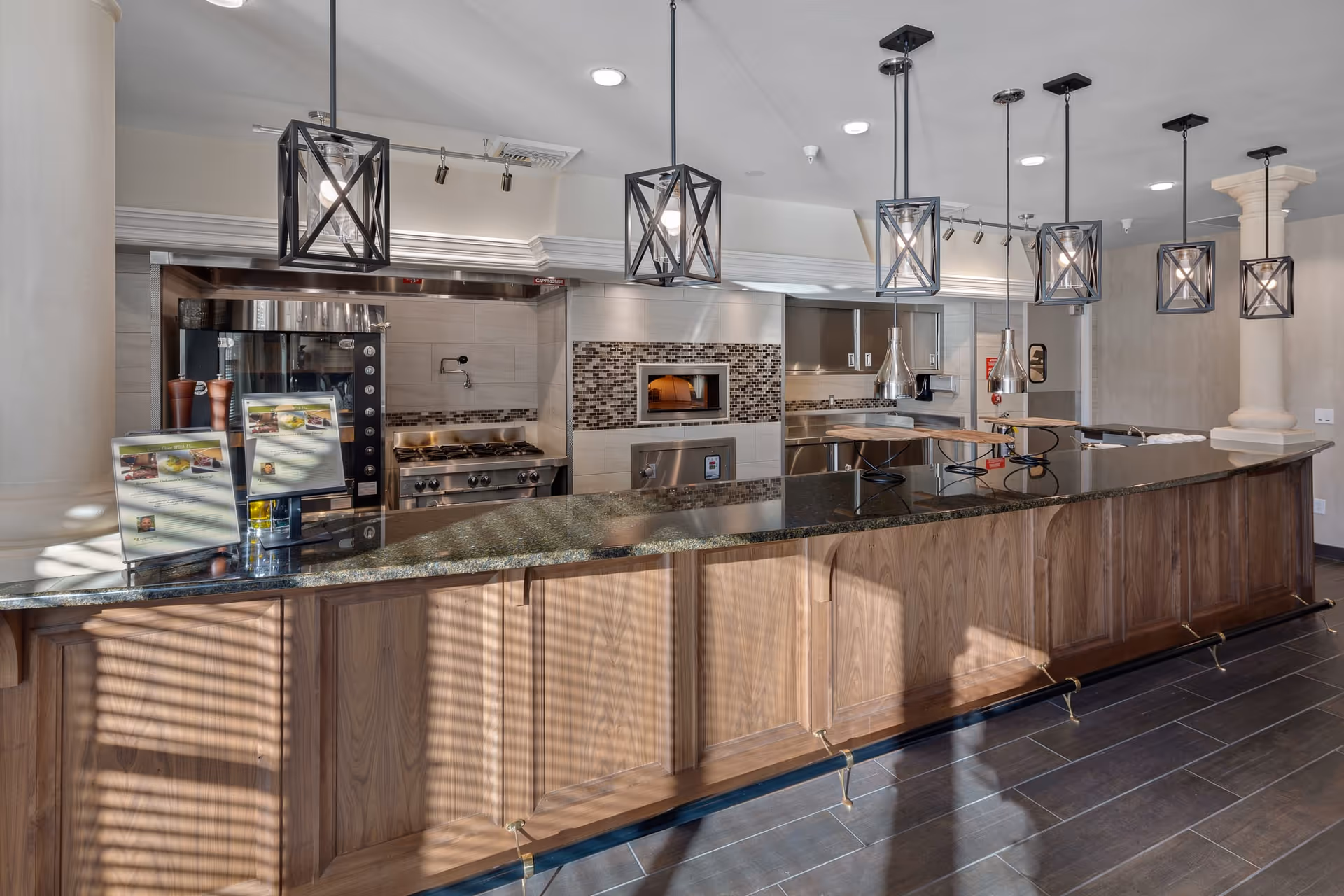 Modern kitchen area with a long granite countertop and wooden cabinetry. Above the counter, there are six pendant lights with geometric black metal frames. The kitchen features a stainless steel stove, oven, and a pizza oven embedded in a tiled wall with mosaic accents. The floor is dark wood, and the space is well-lit with natural light casting shadows through window blinds.