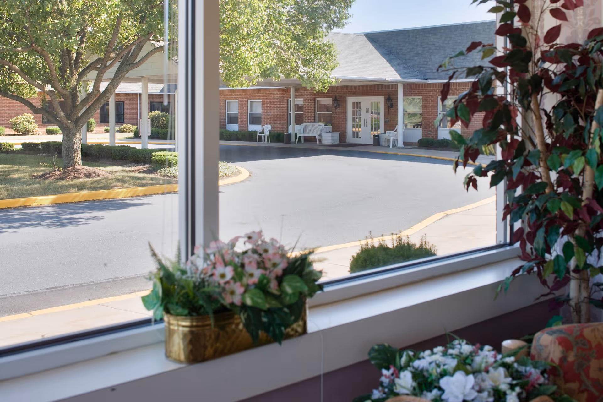 View through a large window showing the exterior entrance of a brick building with a covered drop-off area, white chairs, and a tree with green leaves. Inside, there are potted plants and flowers on the windowsill and nearby furniture.