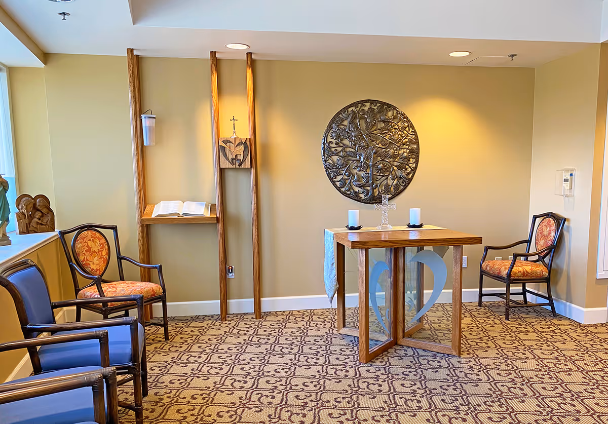A small, quiet room with a religious theme featuring a wooden table with a cross and two candles on top, surrounded by four chairs with patterned upholstery. On the wall behind the table is a large circular metal artwork depicting a tree. To the left, there is a wooden stand holding an open book and a small cross, with a statue on the windowsill.