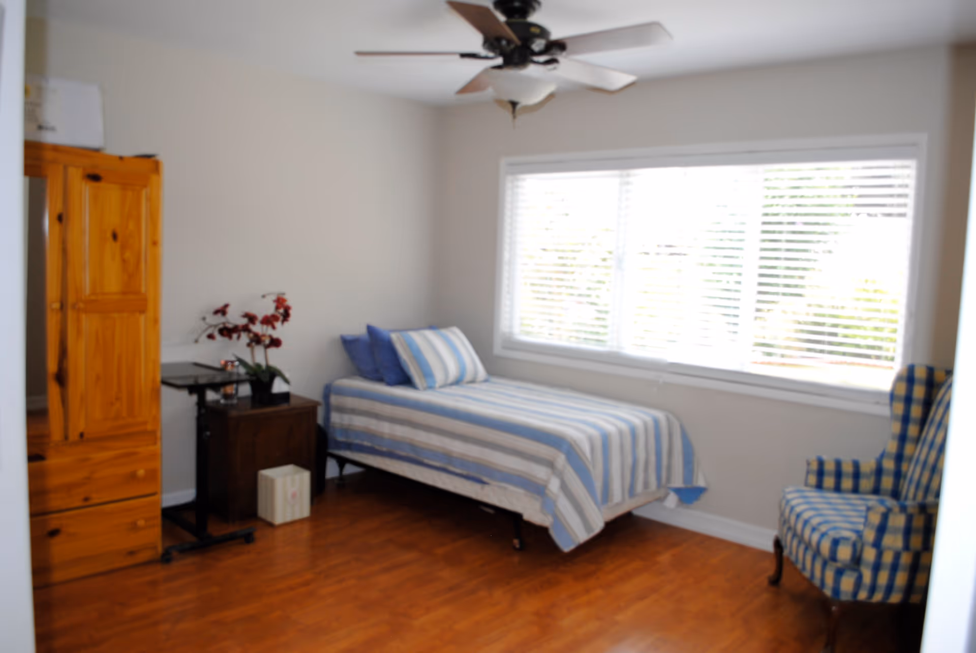 A simple bedroom with a single bed covered in a blue and white striped bedspread, two blue pillows, a wooden wardrobe, a small dark wooden nightstand with a plant on it, a small rolling table, a blue and yellow checkered armchair, and a large window with white blinds letting in natural light. The floor is wooden and there is a ceiling fan with light above.
