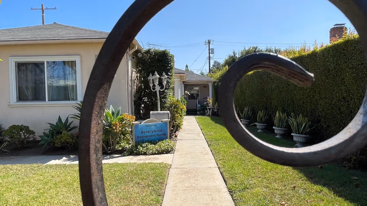 View through a decorative iron gate of a walkway leading to a small beige retirement home with a 'Beverlywood Retirement Home' sign and manicured lawn.
