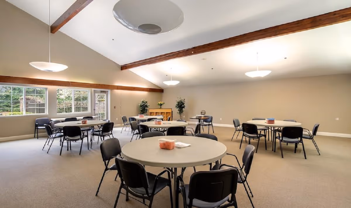 A spacious room with several round tables and black chairs arranged around them. The room has beige walls, a high ceiling with wooden beams, and large windows letting in natural light. There are a few potted plants and a piano against the far wall.