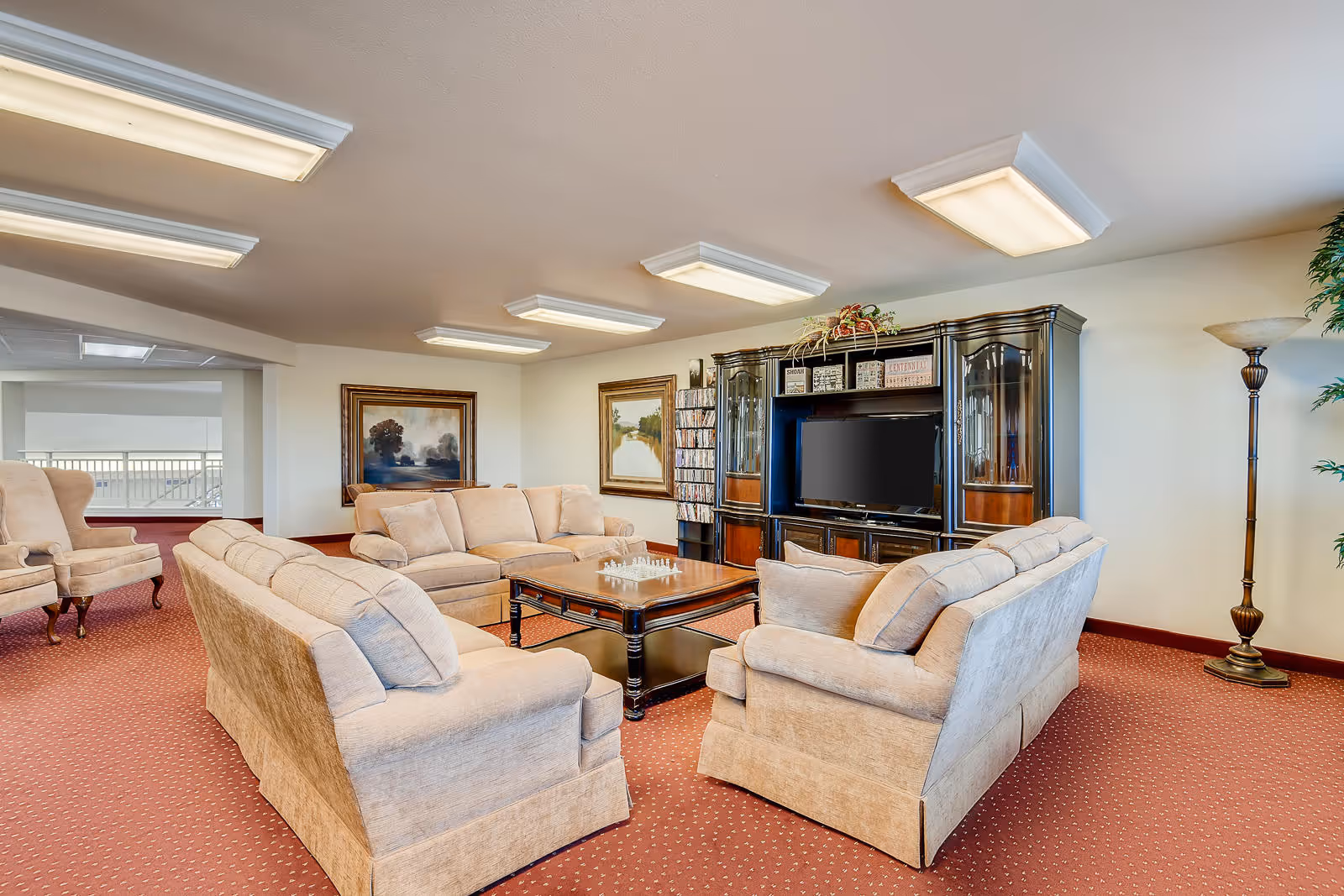 A spacious living room area with beige upholstered sofas arranged around a wooden coffee table with a chessboard on it. The room has a large dark wood entertainment center with a flat-screen TV and decorative items. There are two framed landscape paintings on the walls, a tall floor lamp, and a plant in the corner. The carpet is red with a subtle pattern, and the ceiling has multiple rectangular fluorescent light fixtures.