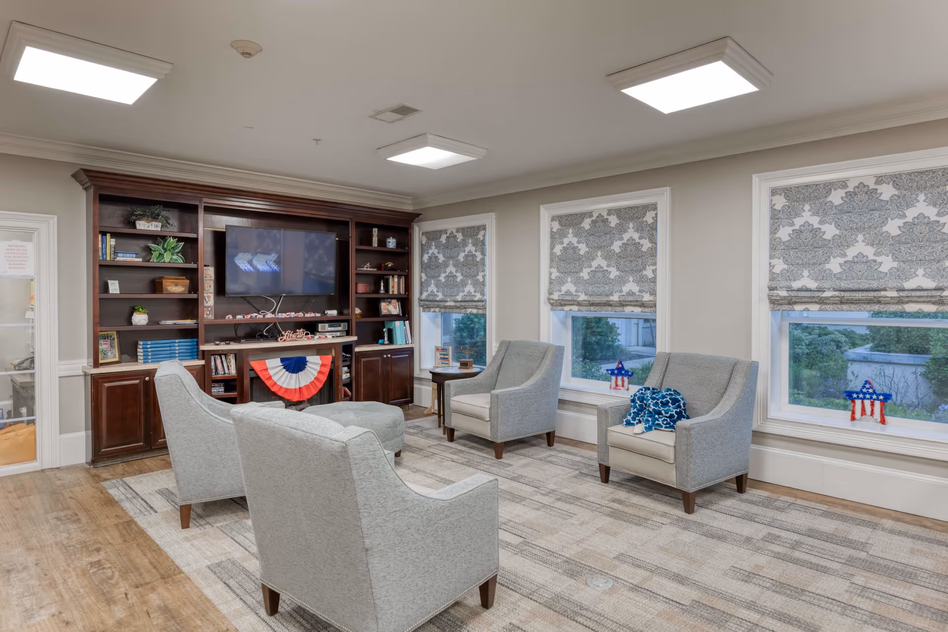 A cozy living room area with four gray upholstered armchairs arranged around a matching ottoman on a patterned rug. Behind the chairs is a large wooden entertainment center with shelves holding books, plants, and decorative items, and a flat-screen TV mounted in the center. The room has three windows with patterned Roman shades and small patriotic star decorations on the windowsills. The floor is wood with a light-colored rug, and the ceiling has recessed lighting.