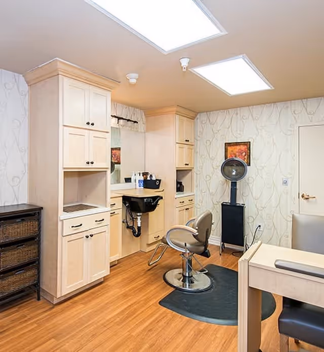 Interior view of a salon area in a senior living facility with a styling chair on a black mat, a hair washing sink, light wood cabinetry, a hair dryer on a stand, and a small table with a chair. The room has wood flooring and patterned wallpaper.