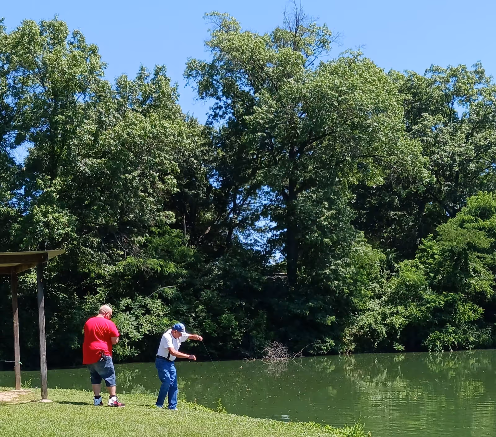 Two people fishing at the edge of a tree-lined pond on a sunny day.