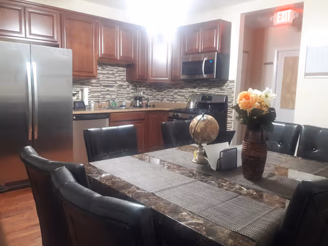 Interior view of a kitchen and dining area featuring dark wooden cabinets, a stainless steel refrigerator, stove, and microwave. A dining table with a marble-like surface is surrounded by black cushioned chairs. On the table, there is a vase with flowers, a small globe, and a napkin holder. An exit sign is visible above a door in the background.