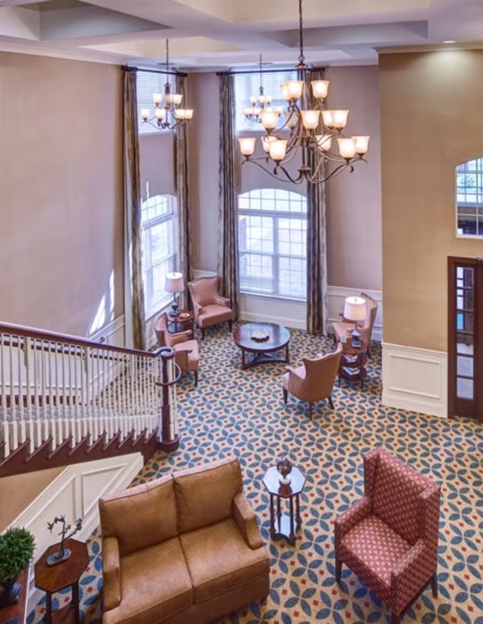 A spacious and well-lit living room area with high ceilings, large windows with long curtains, and multiple chandeliers. The room features a patterned carpet, a brown sofa, a red patterned armchair, several pink armchairs arranged around a round wooden coffee table, and side tables with lamps. A staircase with white spindles and a wooden handrail is visible on the left side.