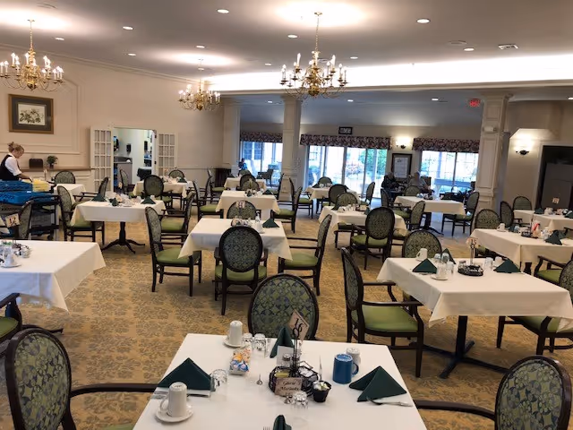 A spacious dining room in a senior living facility with multiple tables covered in white tablecloths, each set with green folded napkins, cups, and condiments. The room features patterned carpet, chandeliers hanging from the ceiling, and large windows letting in natural light. A staff member is seen attending to one of the tables.