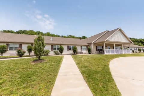 Single-story beige skilled nursing building with a covered entrance, paved walkway, and lawn under a clear blue sky.