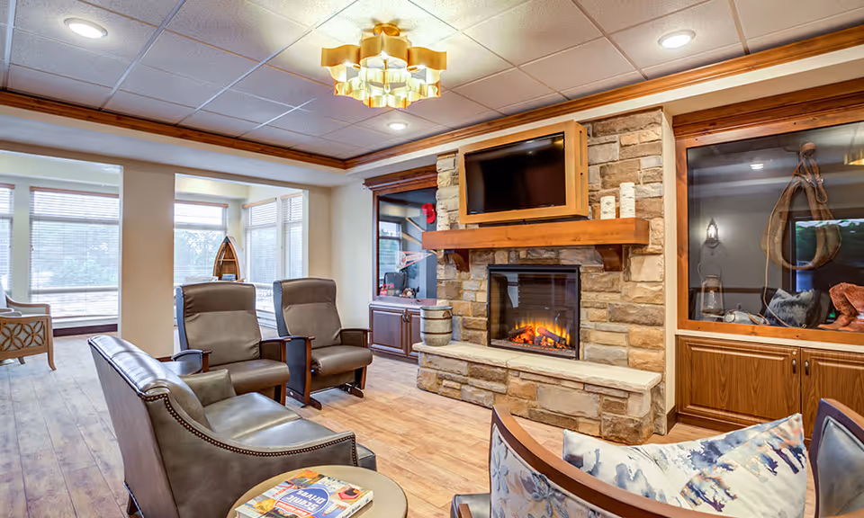 A cozy living room area with a stone fireplace, a mounted flat-screen TV above it, and comfortable seating including leather chairs and a sofa. The room features wood accents, large windows letting in natural light, and a decorative ceiling light fixture.