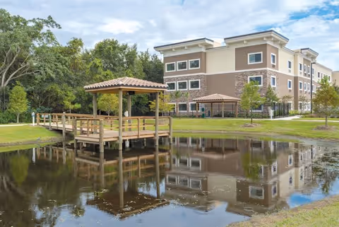 A three-story senior living facility building with beige and brown exterior walls, surrounded by green grass and trees. In the foreground, there is a wooden dock with a small covered gazebo extending over a calm pond that reflects the building and dock. The sky is partly cloudy.