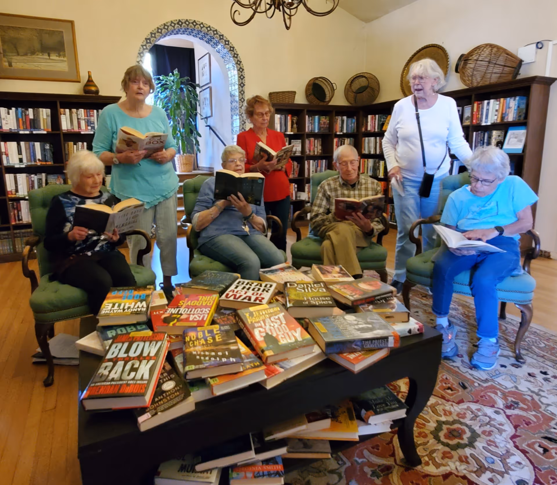 A group of elderly residents reading books together in a cozy library-style common room with bookshelves and a table piled with books.