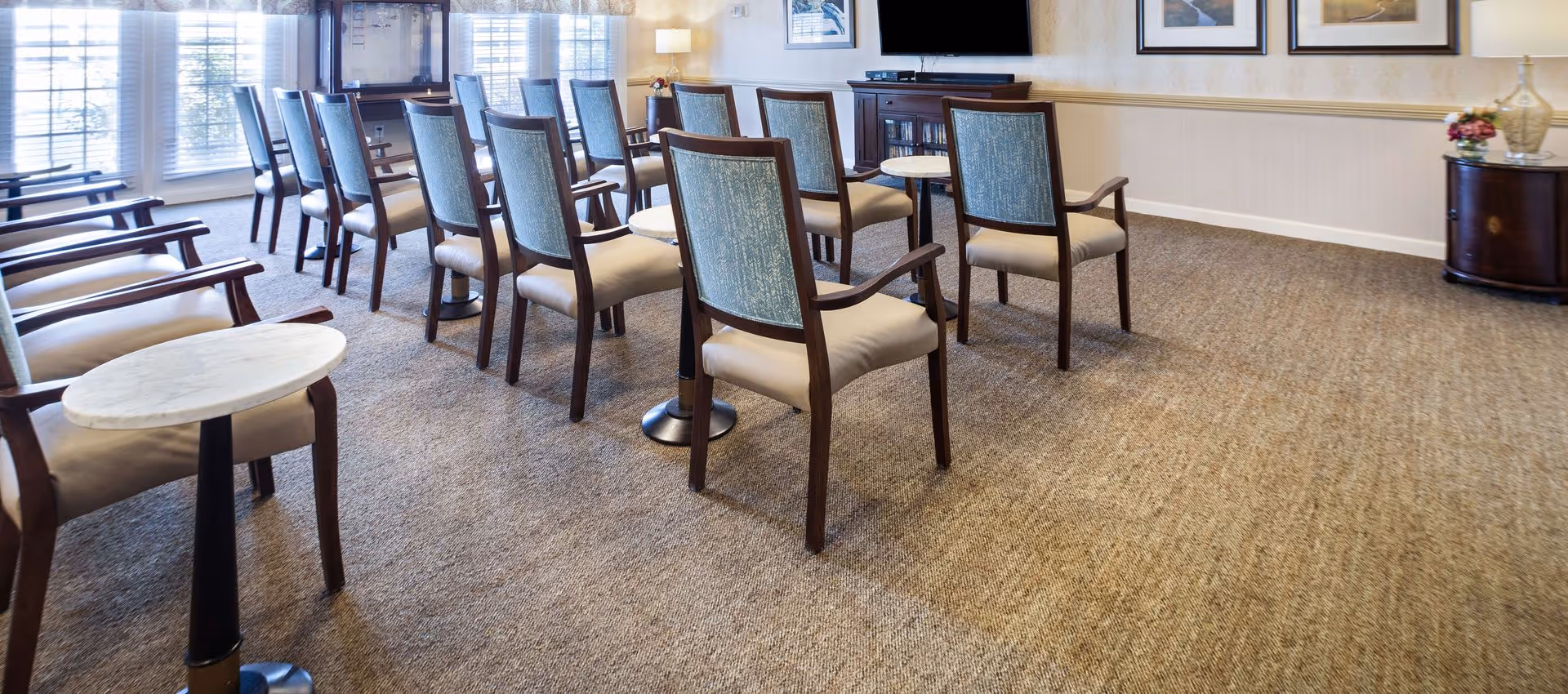 Bright communal activity room with rows of upholstered chairs, small round side tables, a TV console, and large windows.